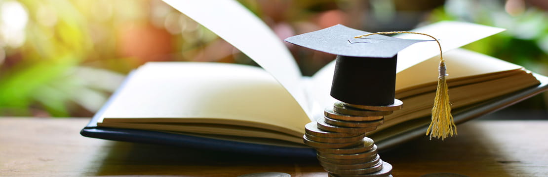 Small paper graduation cap sitting on top of a stack of coins. An open book can be seen the background.
